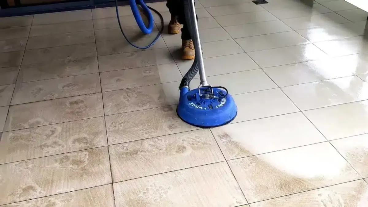 A person cleans a tiled floor with a blue rotary cleaning machine.