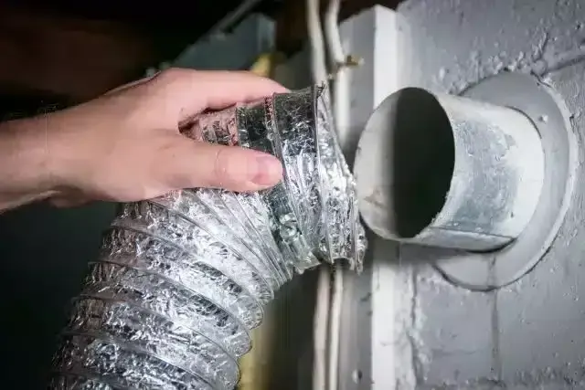 Hand holding flexible dryer vent hose near wall opening.