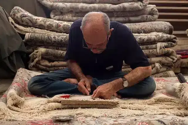 Man working on a rug, surrounded by stacks of rolled rugs.
