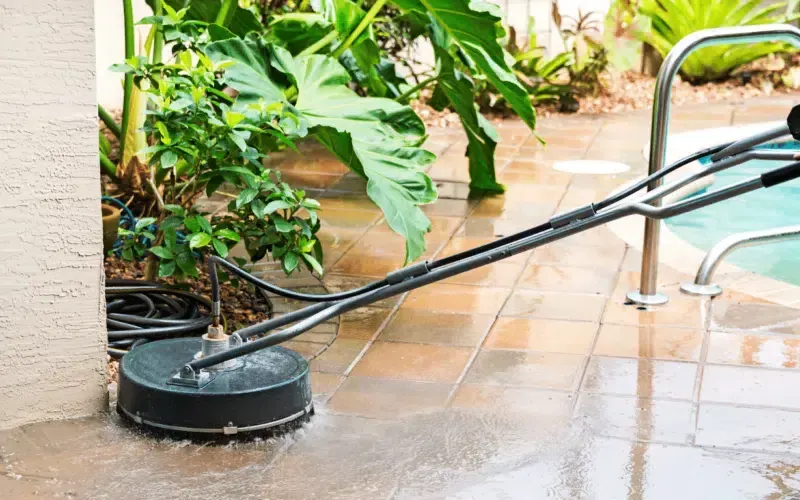 Pressure washer cleaning a tiled patio next to a swimming pool.