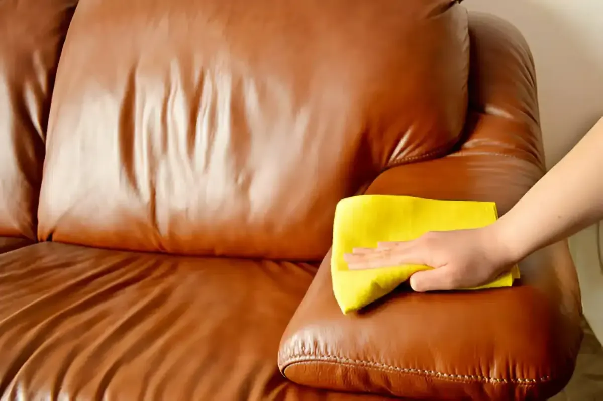 Hand cleaning a brown leather couch with a yellow cloth.