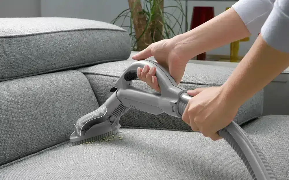 Hands using a cleaning tool on a grey fabric couch cushion.