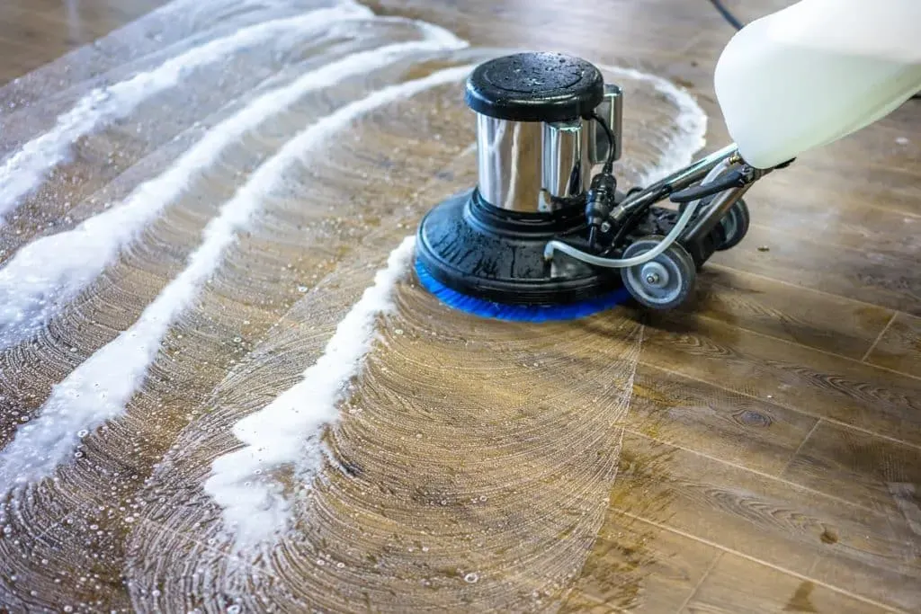 Floor cleaning machine scrubbing a wooden floor with soap and water.