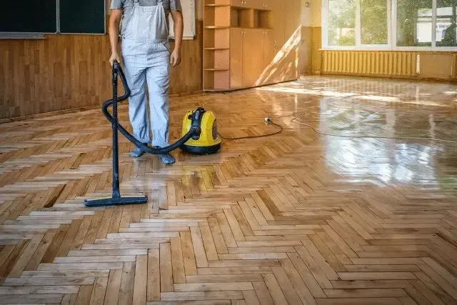 A person in overalls vacuums a wooden parquet floor with a yellow industrial vacuum cleaner.