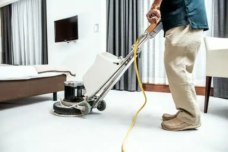Person using a floor buffer machine on a carpeted floor in a room with a bed and curtains.