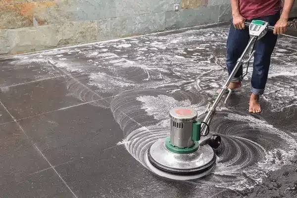 Person cleaning a tiled floor with a floor buffer machine and soap suds.