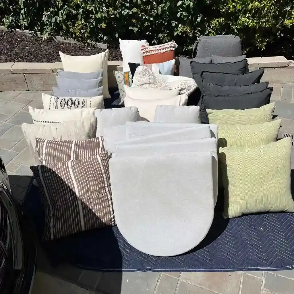 Stack of outdoor cushions and pillows laid out on a blue moving blanket on a paved patio.